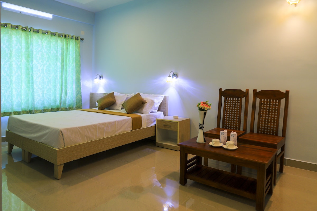 A hotel room with a bed, bedside tables, a nightstand, a wooden chair, and a small table with cups and a vase.