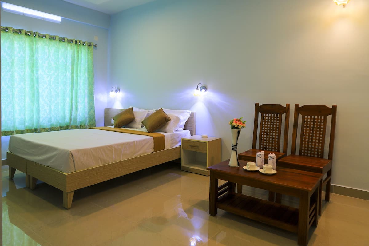 A hotel room with a bed, bedside tables, a nightstand, a wooden chair, and a small table with cups and a vase.