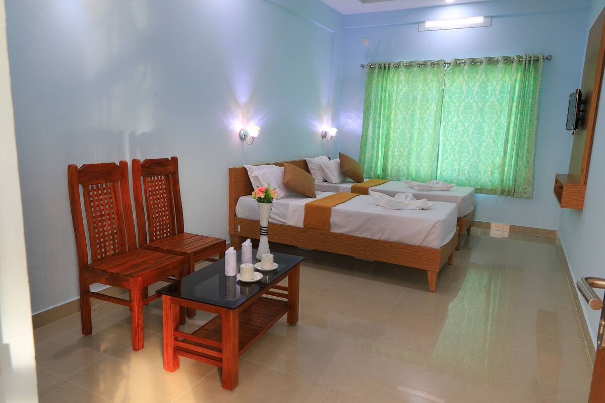 A hotel room with a bed, two wooden chairs, a small table with a vase and candles, and a green curtain.