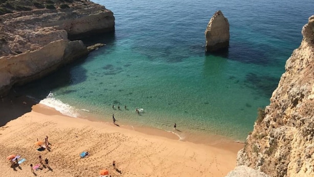 Ein Küstenstrand mit klarem türkisfarbenem Wasser, steilen Klippen und einem Sandufer mit Sonnenschirmen und Menschen.