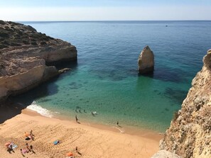 A coastal beach with clear turquoise water, rocky cliffs, and a sandy shore with umbrellas and people.