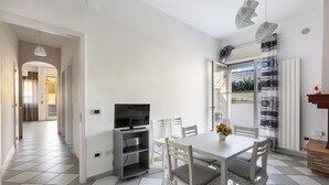 A dining area with a white table and chairs, a television on a stand, and a view through a glass door to the outside.