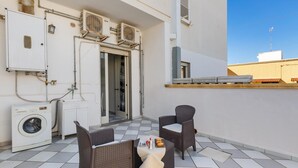 A balcony with a washing machine, two wicker chairs, a small table with a tray of food, and a white cabinet.