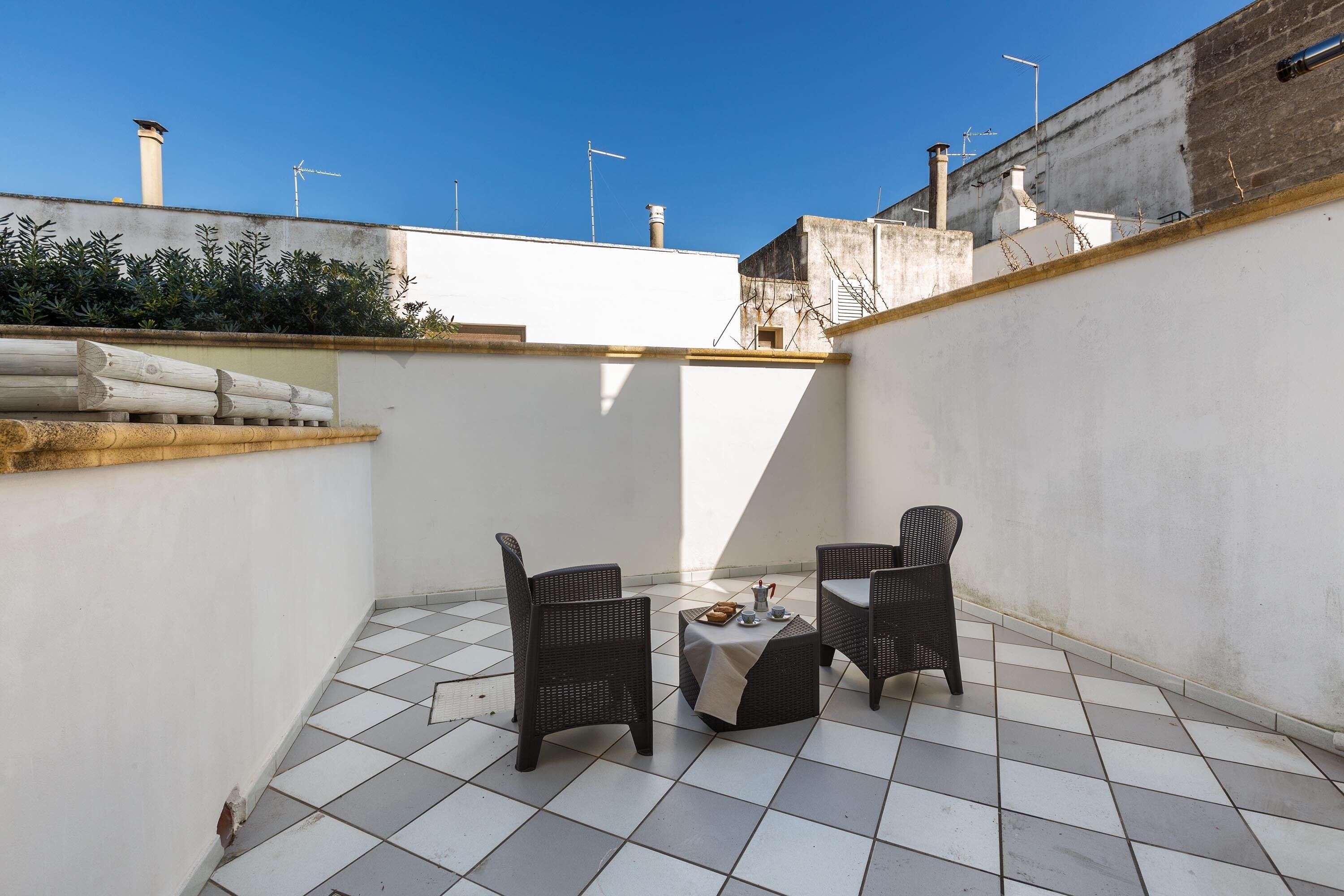A small outdoor patio with two wicker chairs and a small table, surrounded by white tiled flooring and a low white wall.