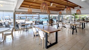 Un vaste espace de restauration intérieur, doté de tables et de chaises en bois, et de grandes fenêtres offrant une vue sur la mer et les bateaux.