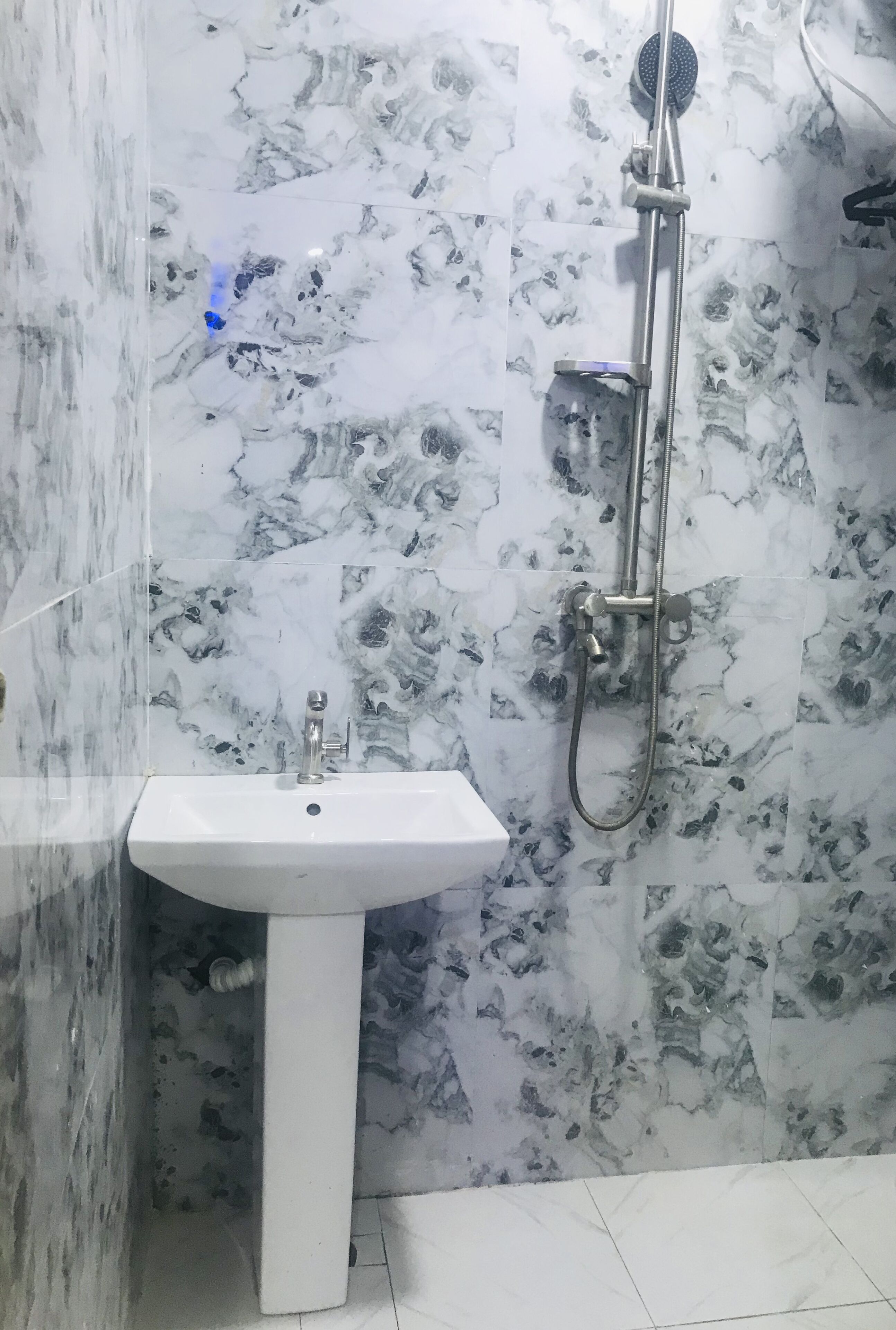 Un baño con pared de diseño de mármol, un lavamanos blanco de pedestal y un cabezal de ducha de mano.
