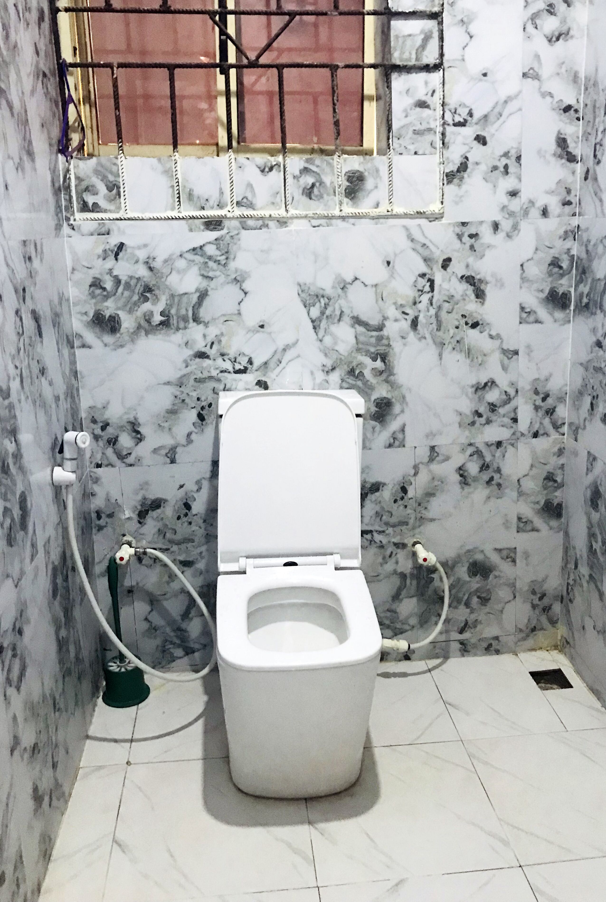 Une salle de bain avec des toilettes, des murs en marbre et une fenêtre grillagée.
