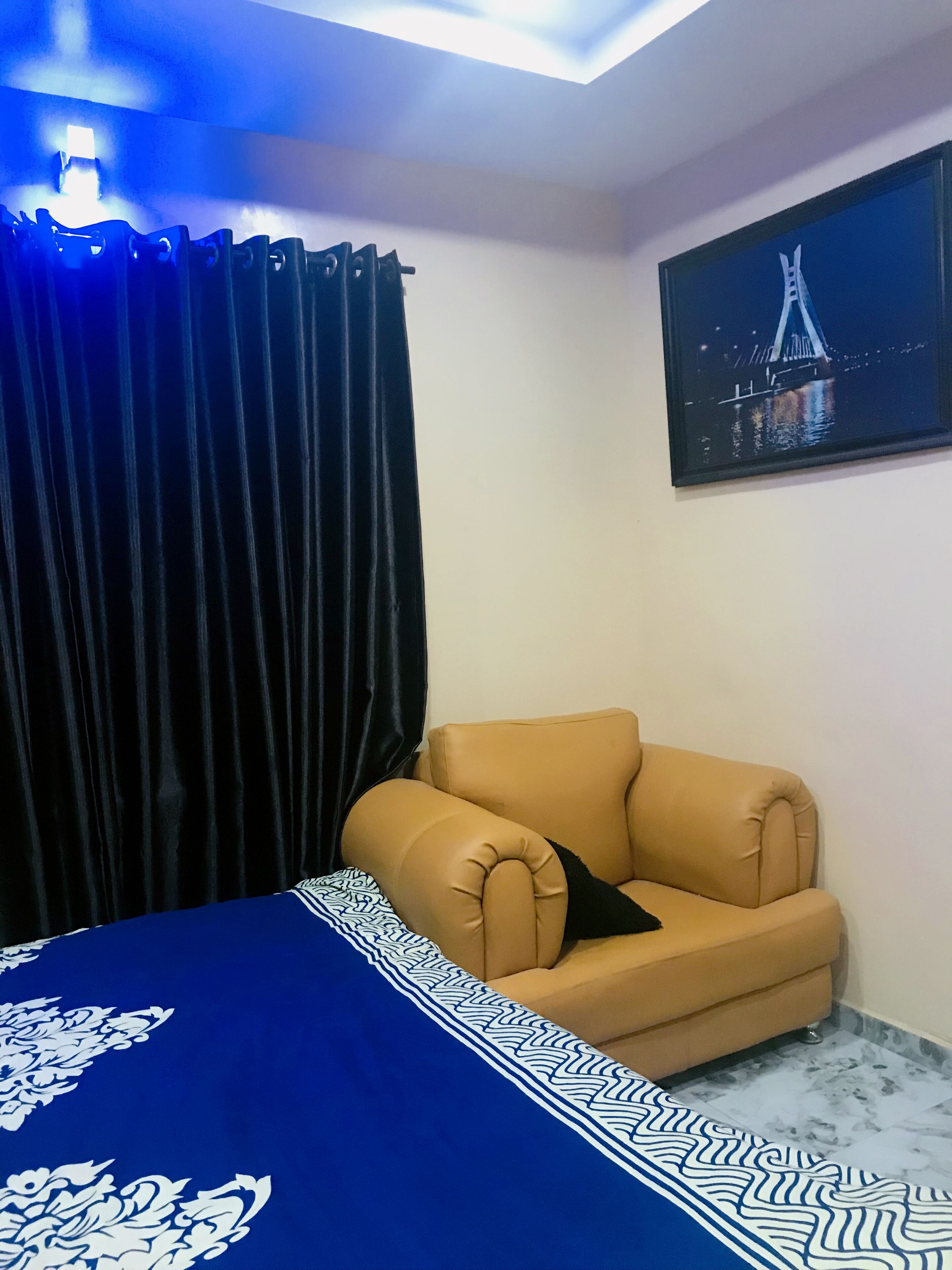 Una habitación con una cama, un sillón color arena, una cortina oscura, un cuadro de un puente y una colcha con estampado.