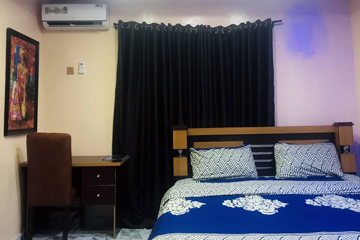 A bedroom with a bed, a desk, an air conditioner, and a curtain.