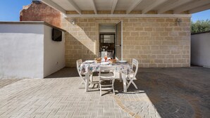 Une terrasse extérieure avec une table dressée pour les repas, des murs en pierre et un espace couvert avec une porte donnant sur la cuisine.