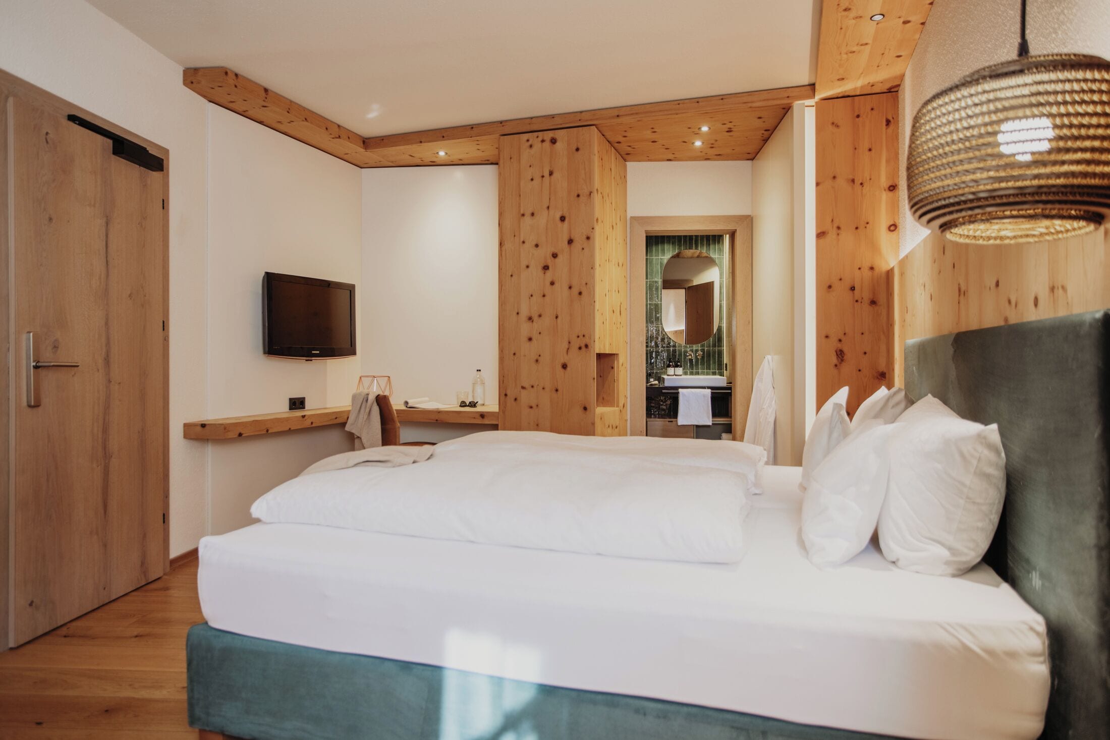 Un dormitorio con una cama grande, vigas de techo de madera, un televisor montado y un baño visible a través de una puerta abierta.