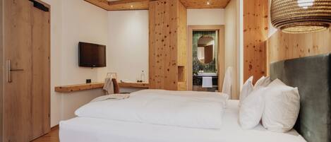 Un dormitorio con una cama grande, vigas de techo de madera, un televisor montado y un baño visible a través de una puerta abierta.