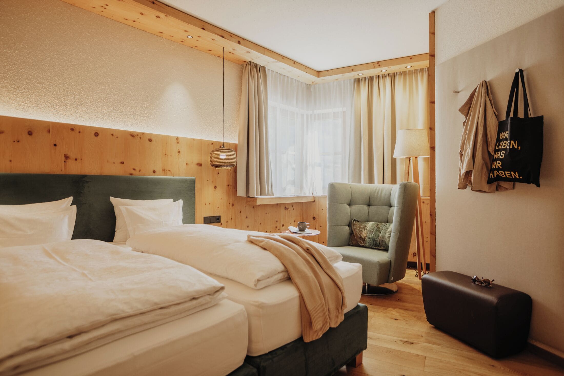 Ein Hotelzimmer mit einem großen Bett, einem kleinen Sessel, einem Nachttisch mit Lampe und einem Koffer.