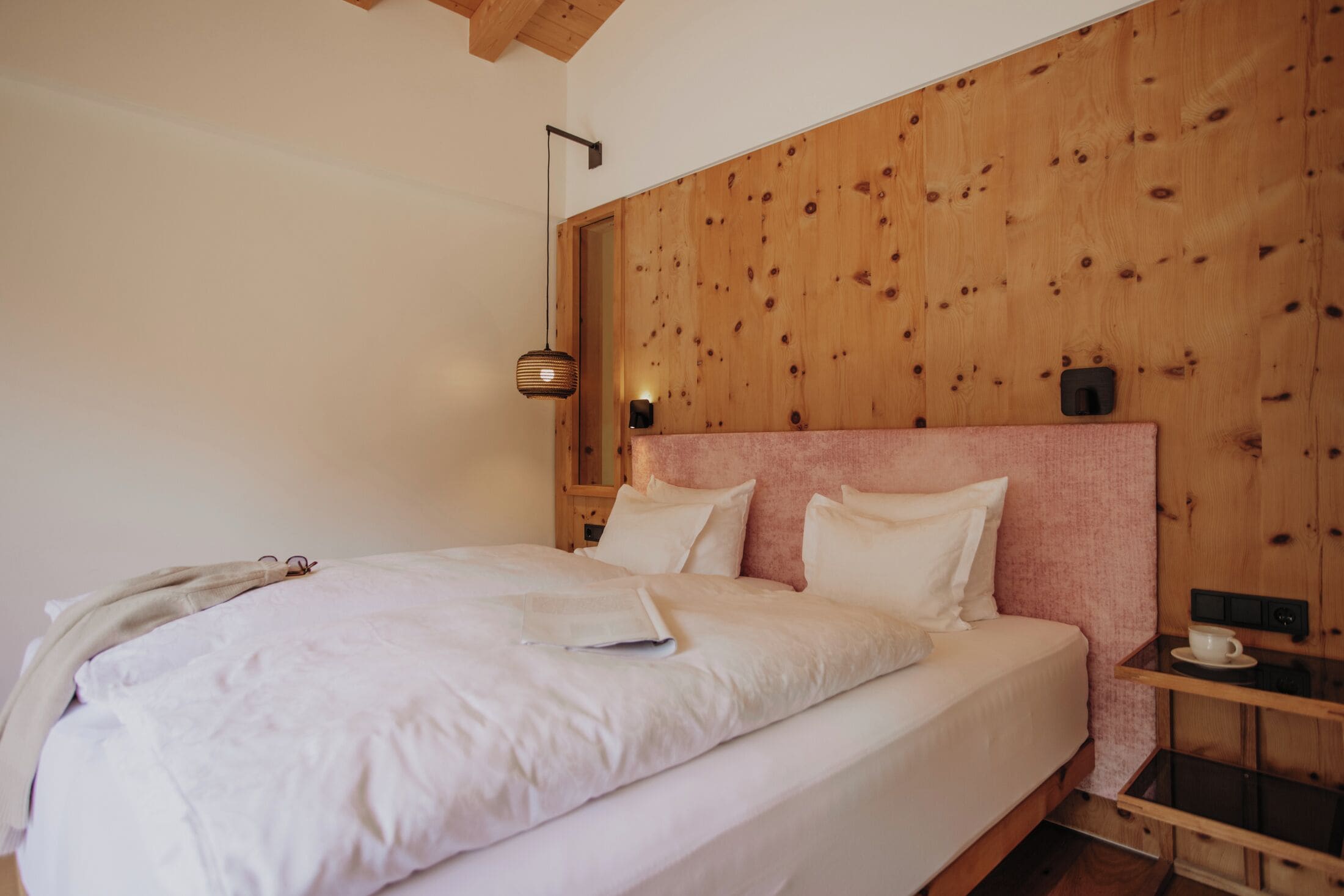 A neatly made bed with white linens, a wooden headboard, and a bedside table with a cup and saucer.