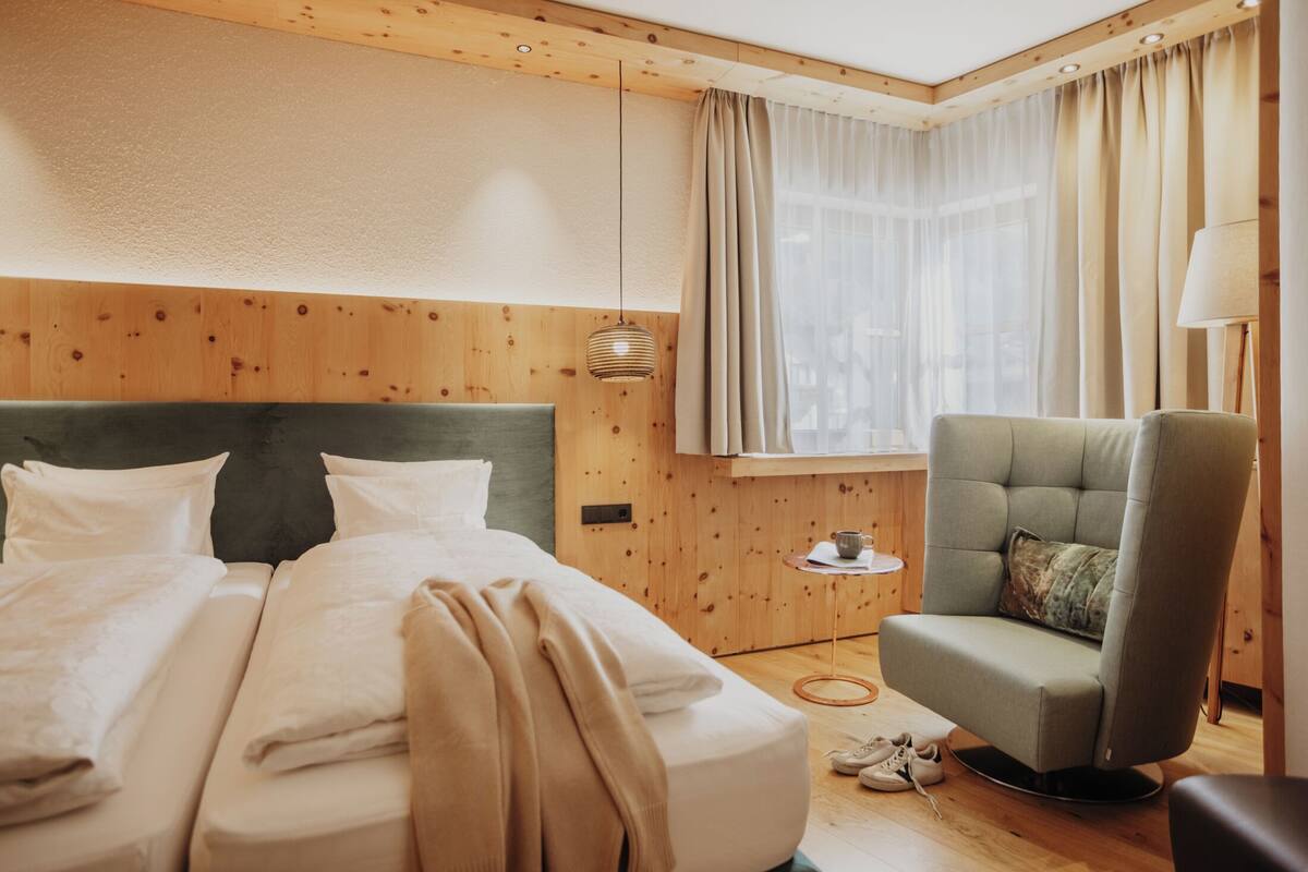 Una habitación de hotel con una cama, una silla, una mesita con una taza y una ventana con cortinas.