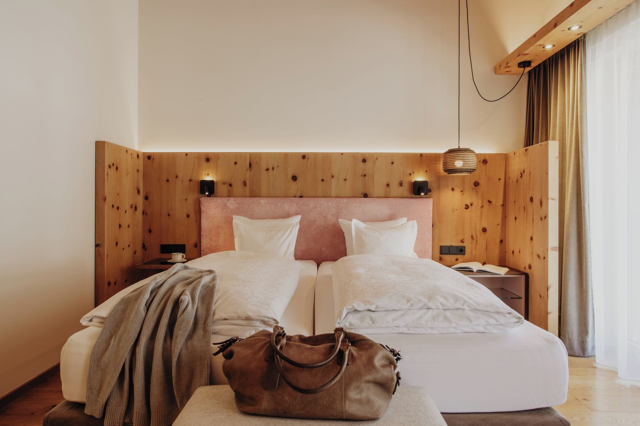 Un lit double avec du linge de lit blanc, une tête de lit en bois, des tables de chevet avec des lampes et un sac à main marron posé sur le lit.