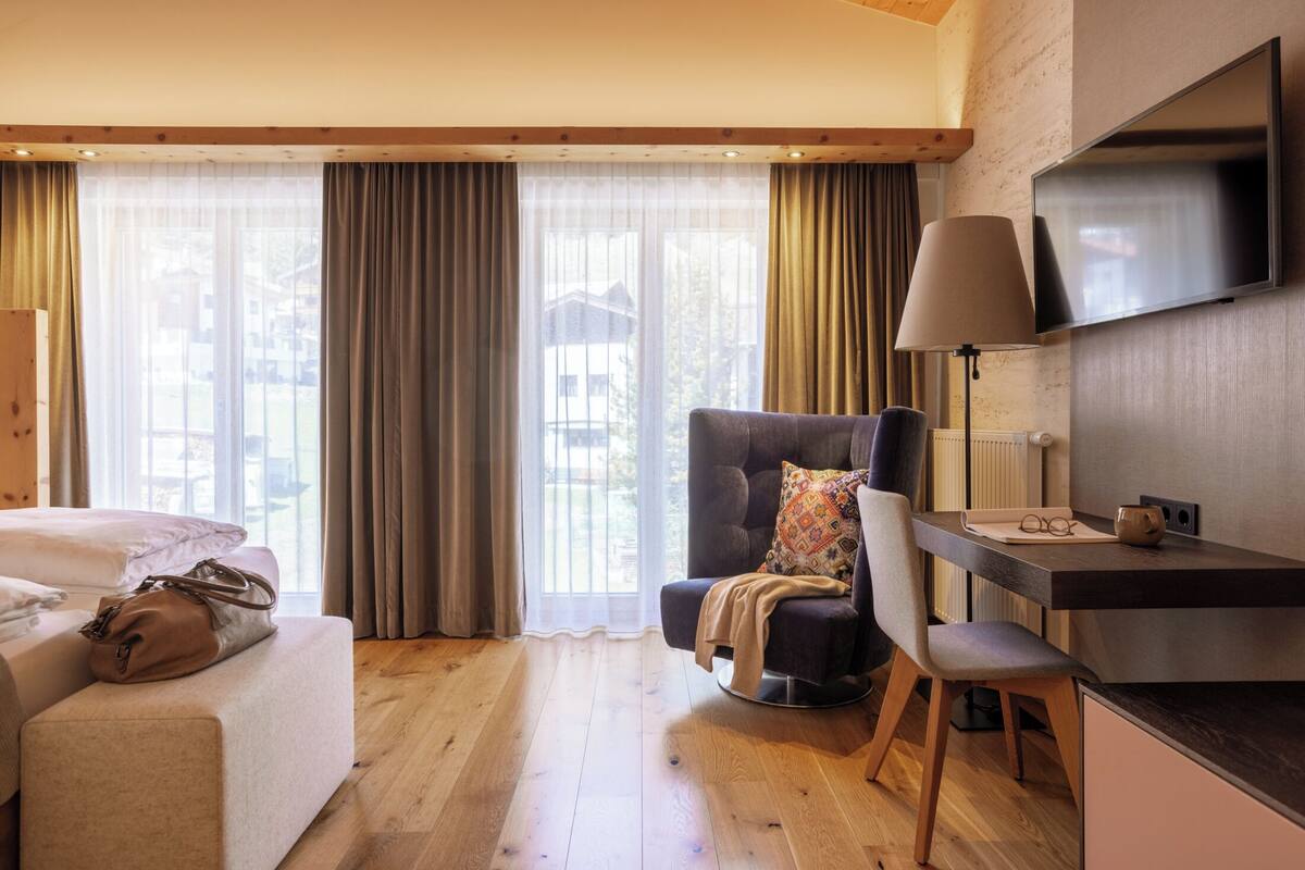 Habitación de hotel con cama, escritorio, silla, televisor y un amplio ventanal con cortinas.
