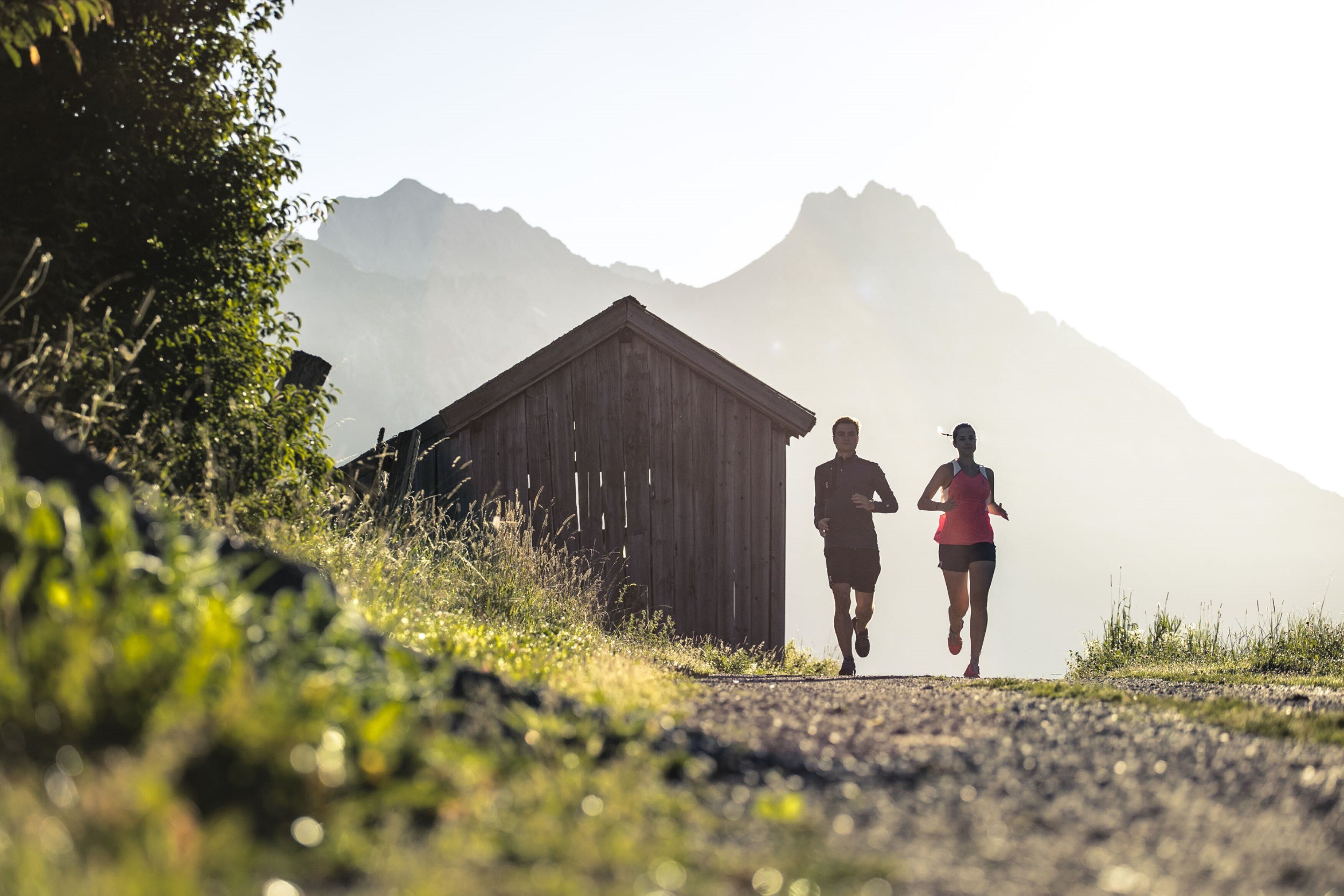 Two people jogging on a gravel path with a wooden structure and mountains in the background.