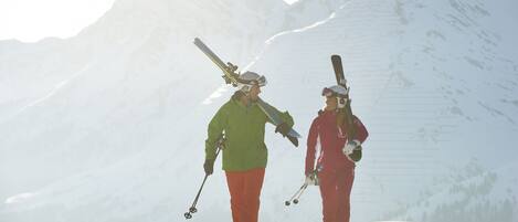 Dos personas caminando sobre una montaña nevada con esquíes, rodeadas de picos cubiertos de nieve.