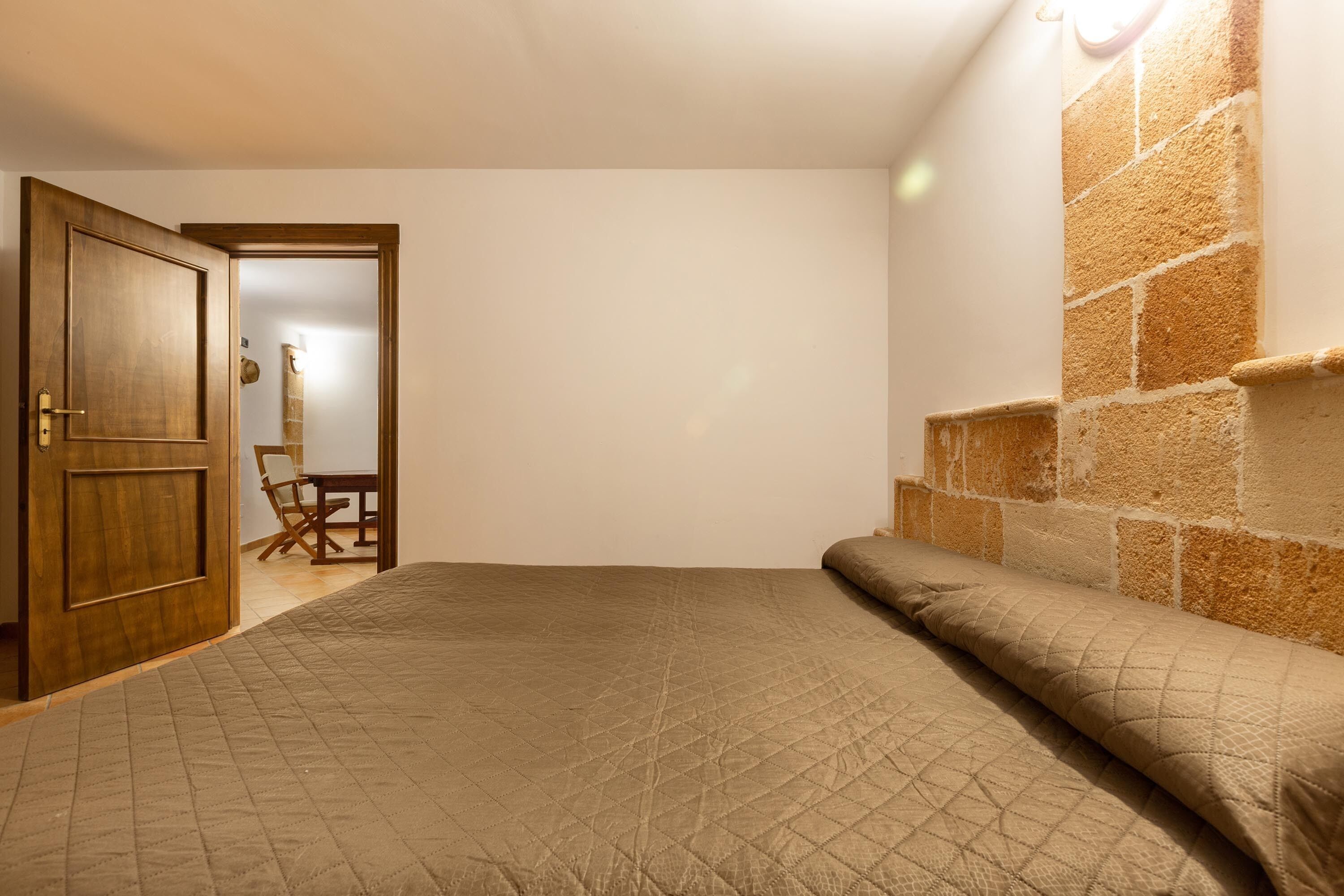 A bedroom with a bed, a wooden door, a stone wall, and a chair in the background.
