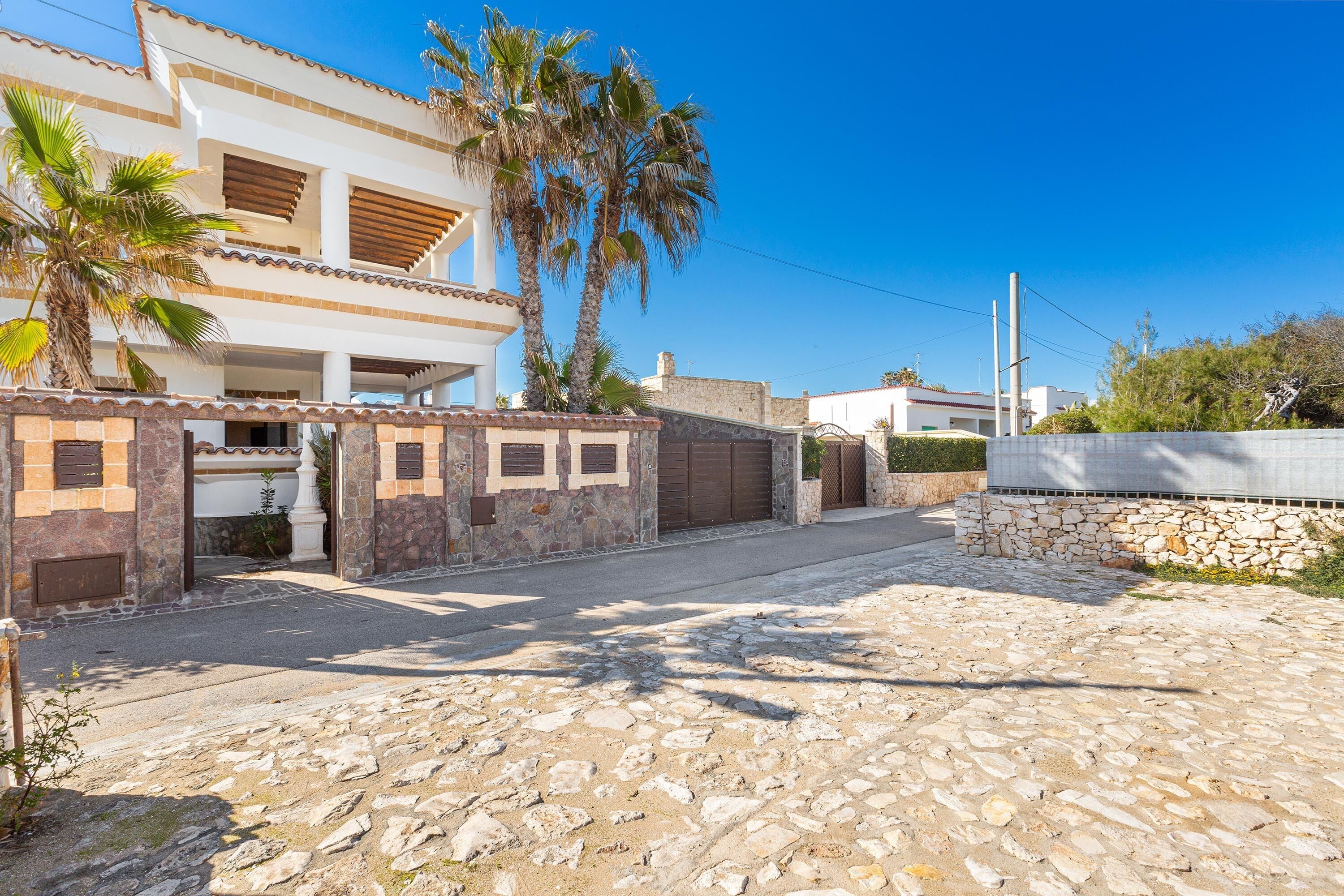 A two-story house with a stone facade, a paved driveway, and palm trees.