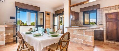 A dining area with a table set for four, surrounded by wicker chairs, a kitchen with a tiled backsplash, and a view through the open door.