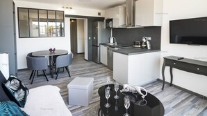 A modern kitchen with a central island, a dining area with a round table and chairs, a living area with a sofa and a coffee table, and a TV mounted on the wall. - Apartment "Irrésistible" (LE CAP D'AGDE)