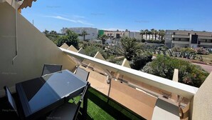 A balcony with a table and chairs, overlooking a landscaped area with palm trees and a building complex in the background. - Apartment "Irrésistible" (LE CAP D'AGDE)