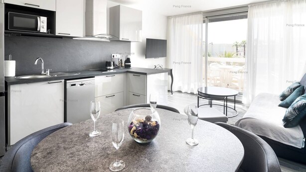 A modern kitchen with a built-in microwave, a coffee machine, and a sink. There is a dining area with a round table and two chairs, and a sofa with cushions. - Apartment "Irrésistible" (LE CAP D'AGDE)
