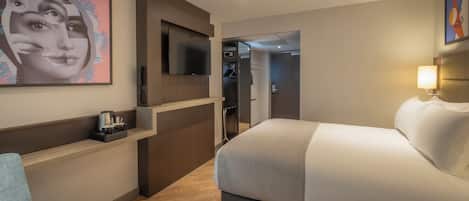 Habitación de hotel con una cama, un televisor montado en la pared, un pequeño rincón de descanso con una silla y un cuadro en la pared.