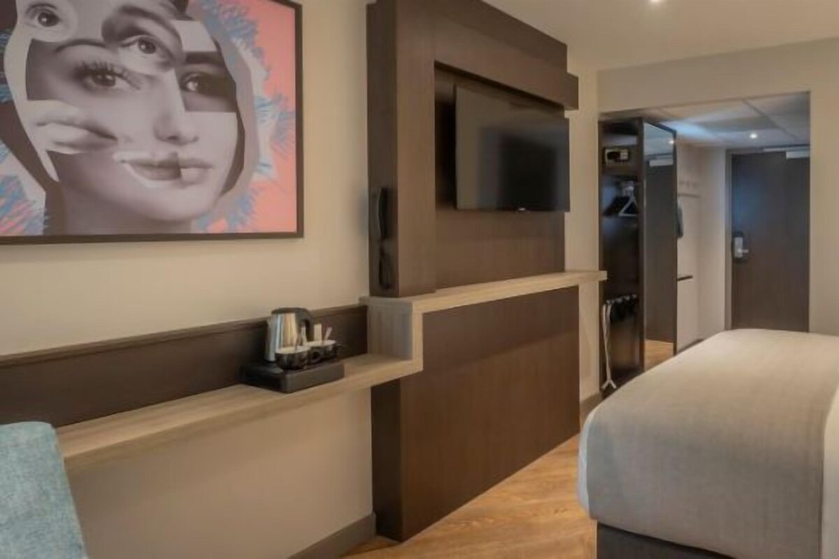 A hotel room with a bed, a TV mounted on the wall, a bedside table with a lamp, and a painting on the wall.