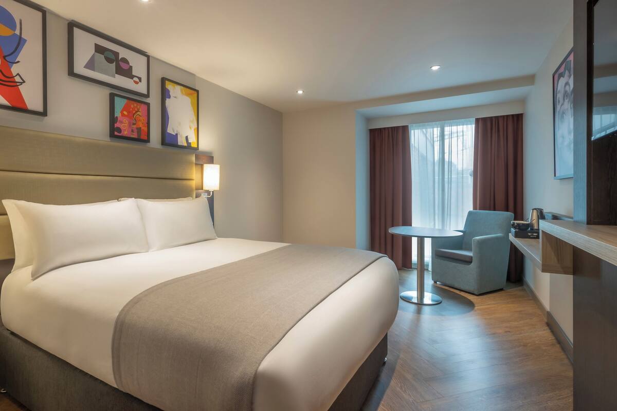 Ein Hotelzimmer mit einem großen Bett, einem kleinen Tisch, einem Stuhl und Bildern an den Wänden.