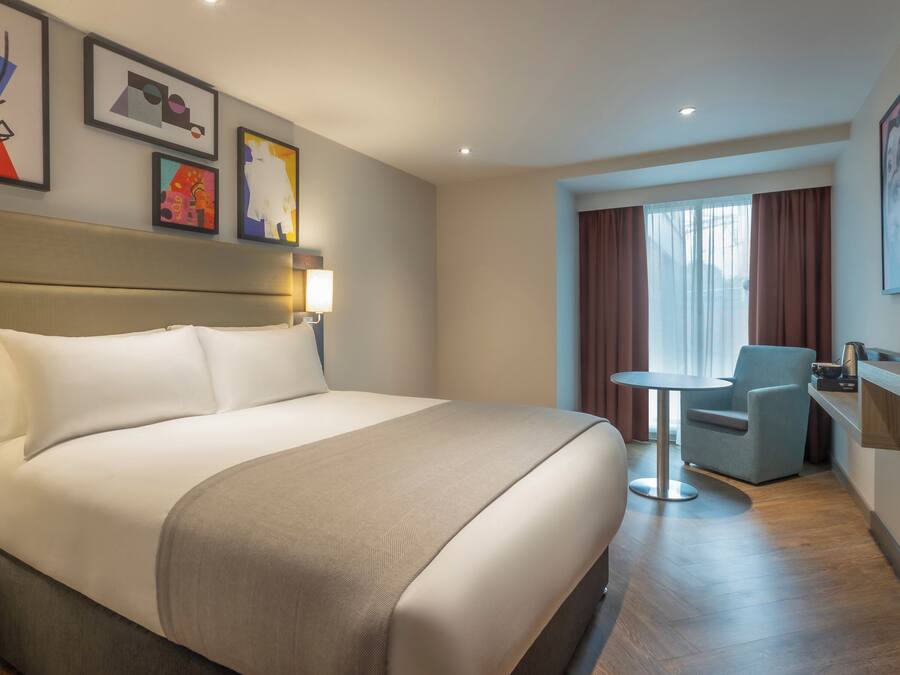 Una habitación de hotel con una cama grande, una mesita pequeña, una silla y obras de arte en las paredes.