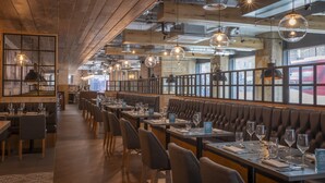 Un restaurante con vigas de madera, luces esféricas colgantes y asientos de cuero. Las mesas están preparadas con copas de vino y menús.