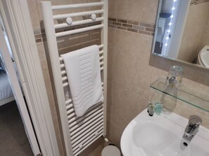 Une salle de bain avec un radiateur blanc, une étagère en verre où sont posés des articles de toilette, et un lavabo avec un miroir au-dessus.