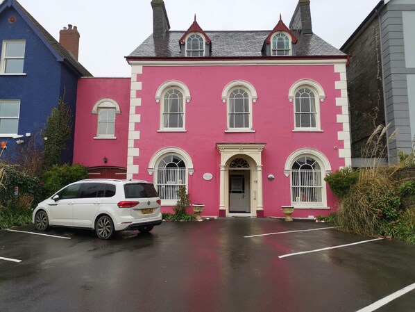 Un bâtiment rose avec une porte et des fenêtres blanches, une voiture blanche garée devant, et un bâtiment bleu sur la gauche.
