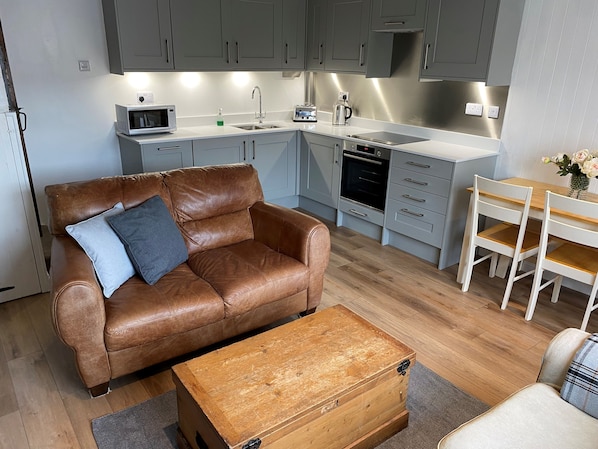 Eine moderne Küche mit grauen Schränken, einer Mikrowelle, einem Wasserkocher und einer Kaffeemaschine. Es gibt ein braunes Ledersofa mit Kissen, eine Holzbank und einen weißen Esstisch mit Stühlen.