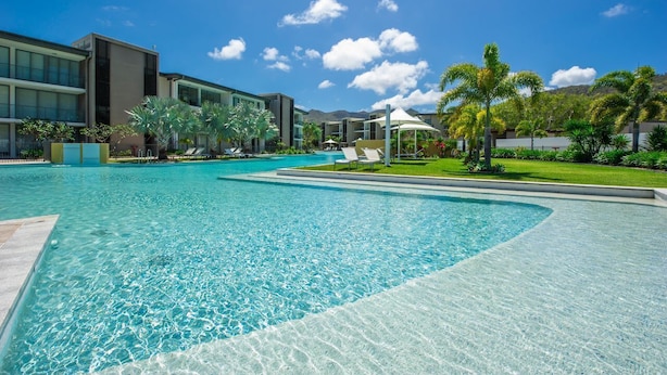 A swimming pool with clear water, surrounded by modern buildings, palm trees, and a clear sky.