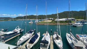 A marina with multiple sailboats docked, a clear sky, and a hilly landscape in the background.