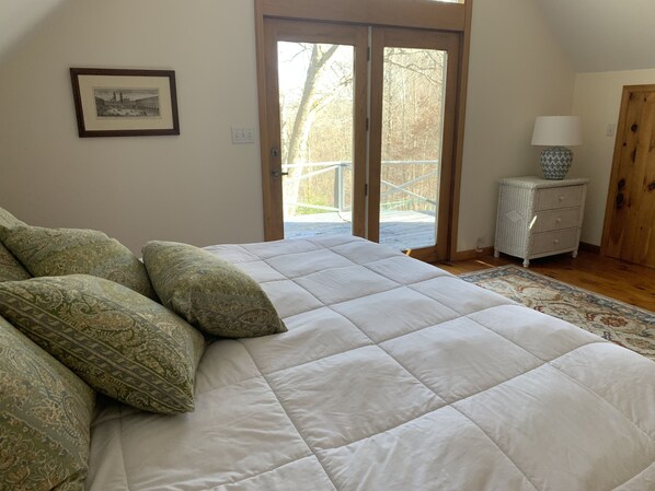 A bedroom with a large bed, patterned pillows, a wooden door, a small bedside table with a lamp, and a framed picture on the wall.