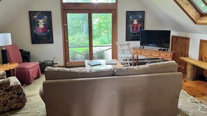Ein Wohnzimmer mit Sofa, Fernseher, Schreibtisch und Stuhl. Zwei Fenster und zwei gerahmte Bilder schmücken die Wände.