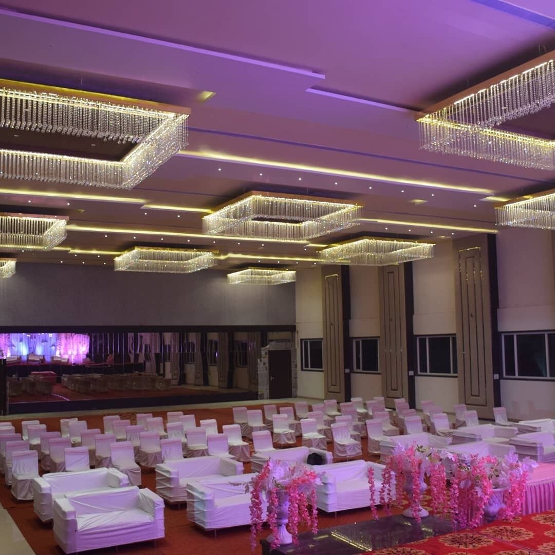 A banquet hall with rows of white chairs, a stage with a purple backdrop, and modern chandeliers.