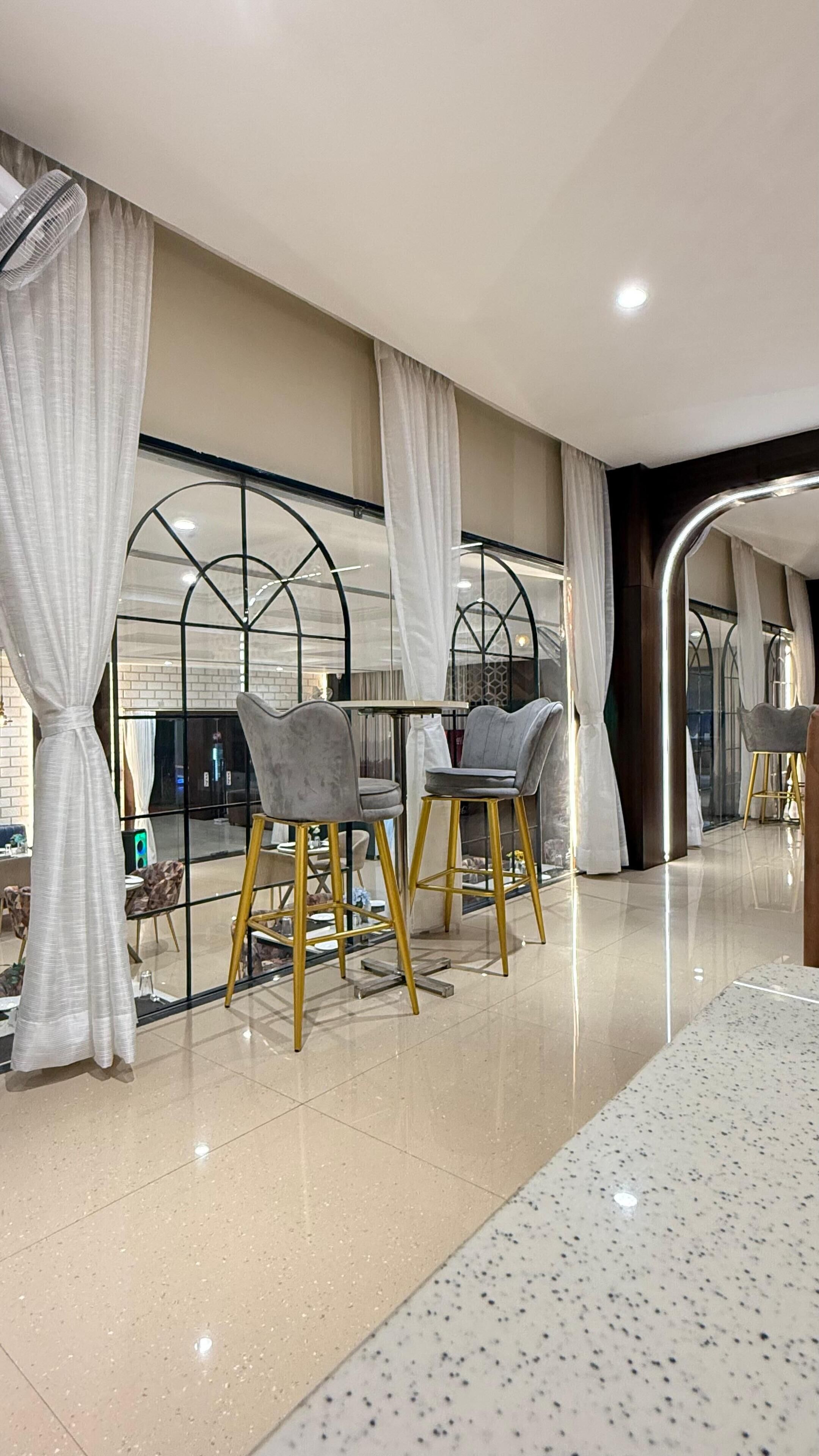 Une chambre d’hôtel moderne avec un grand lit, deux fauteuils aux pieds dorés, une petite table et une grande fenêtre avec des rideaux.