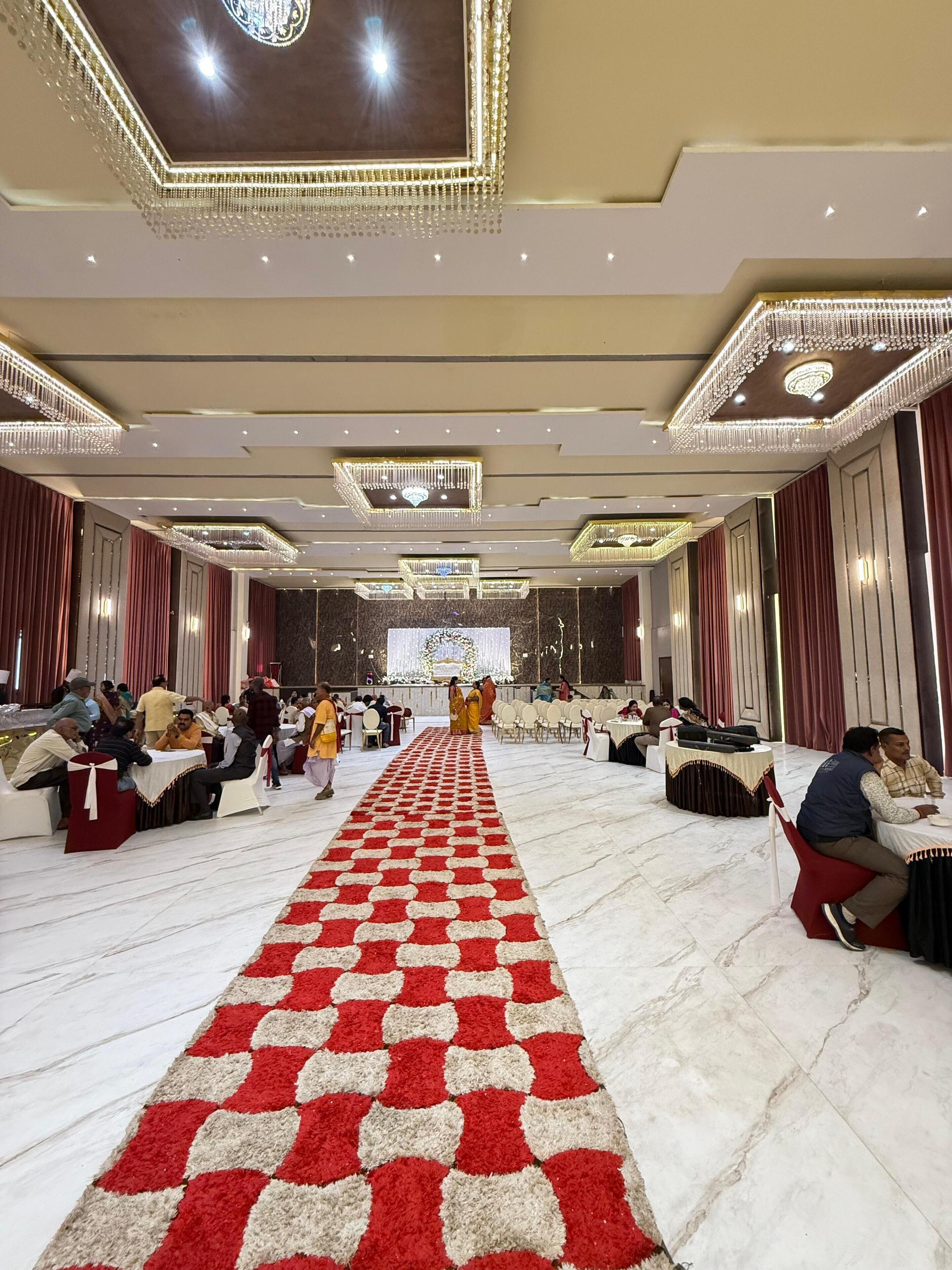 A spacious banquet hall with a red and white checkered carpet leading to a stage, surrounded by tables and chairs.