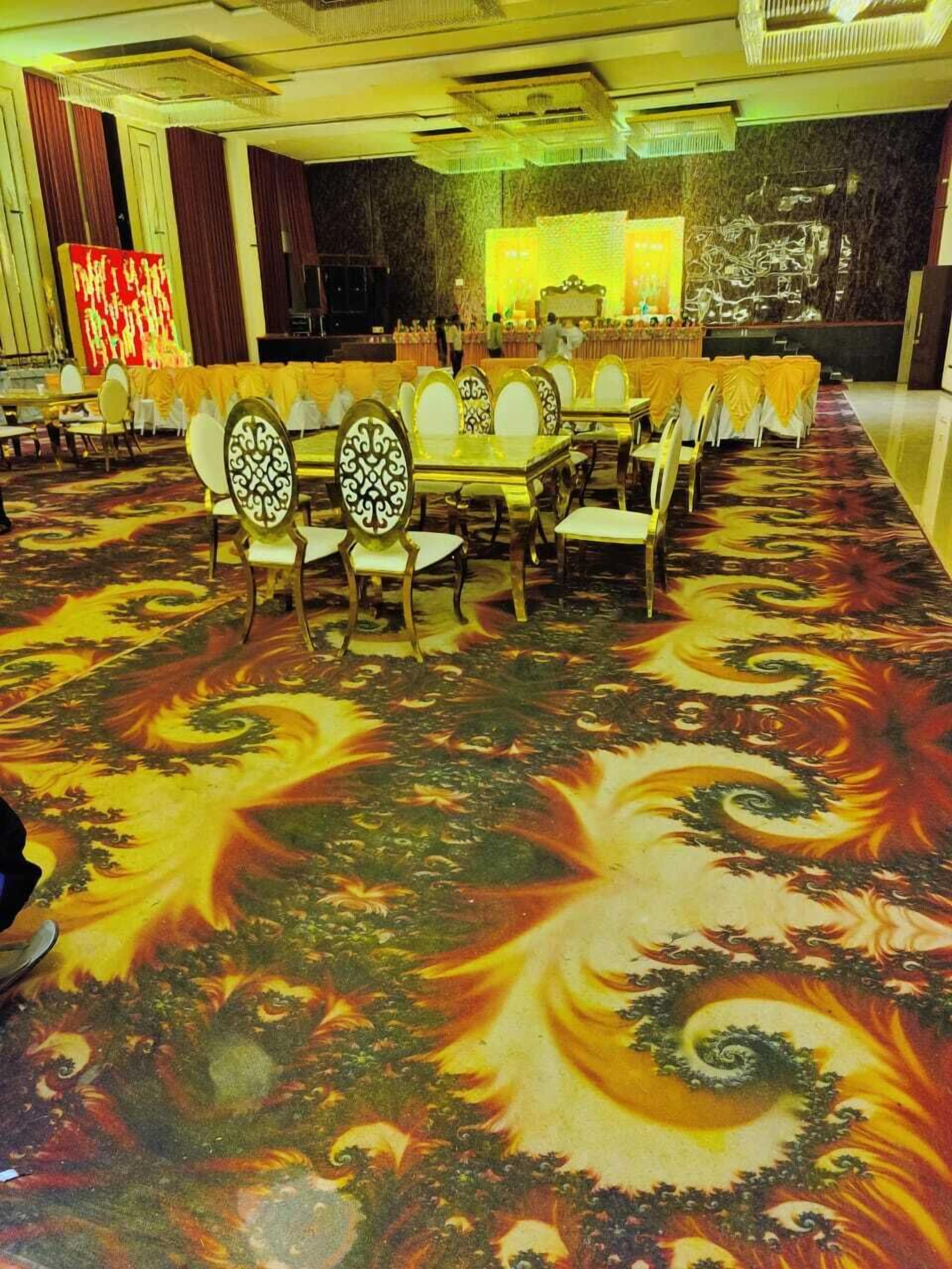 A banquet hall with round tables and chairs, a patterned carpet, and a stage with a red backdrop.