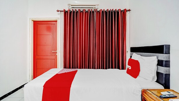 Habitación de hotel con cama, una puerta roja, ventana con cortinas rojas y mesita de noche con teléfono y libro.