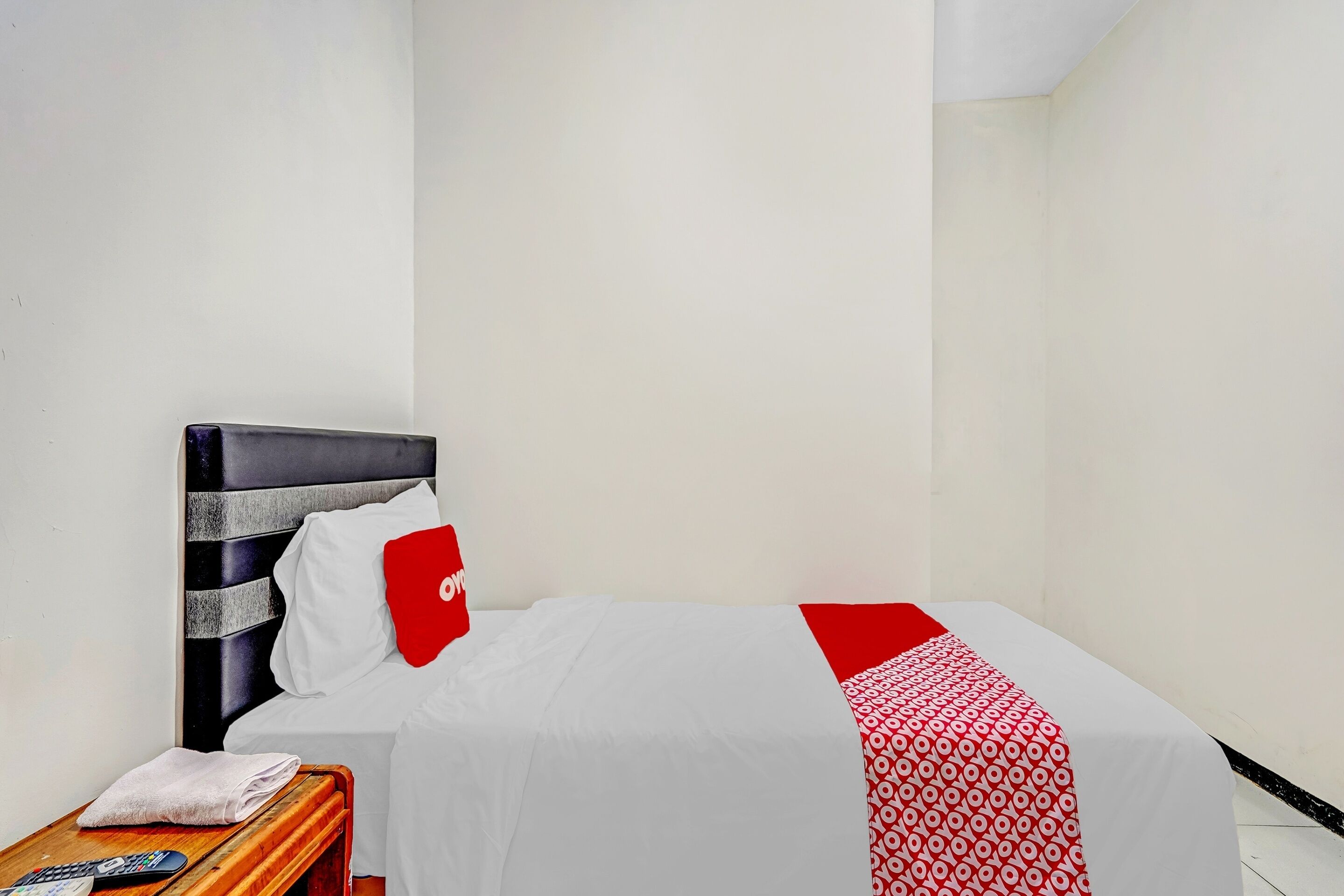 A small, neatly made bed with a white and red bedspread, a wooden nightstand with a remote control, and a red pillow.