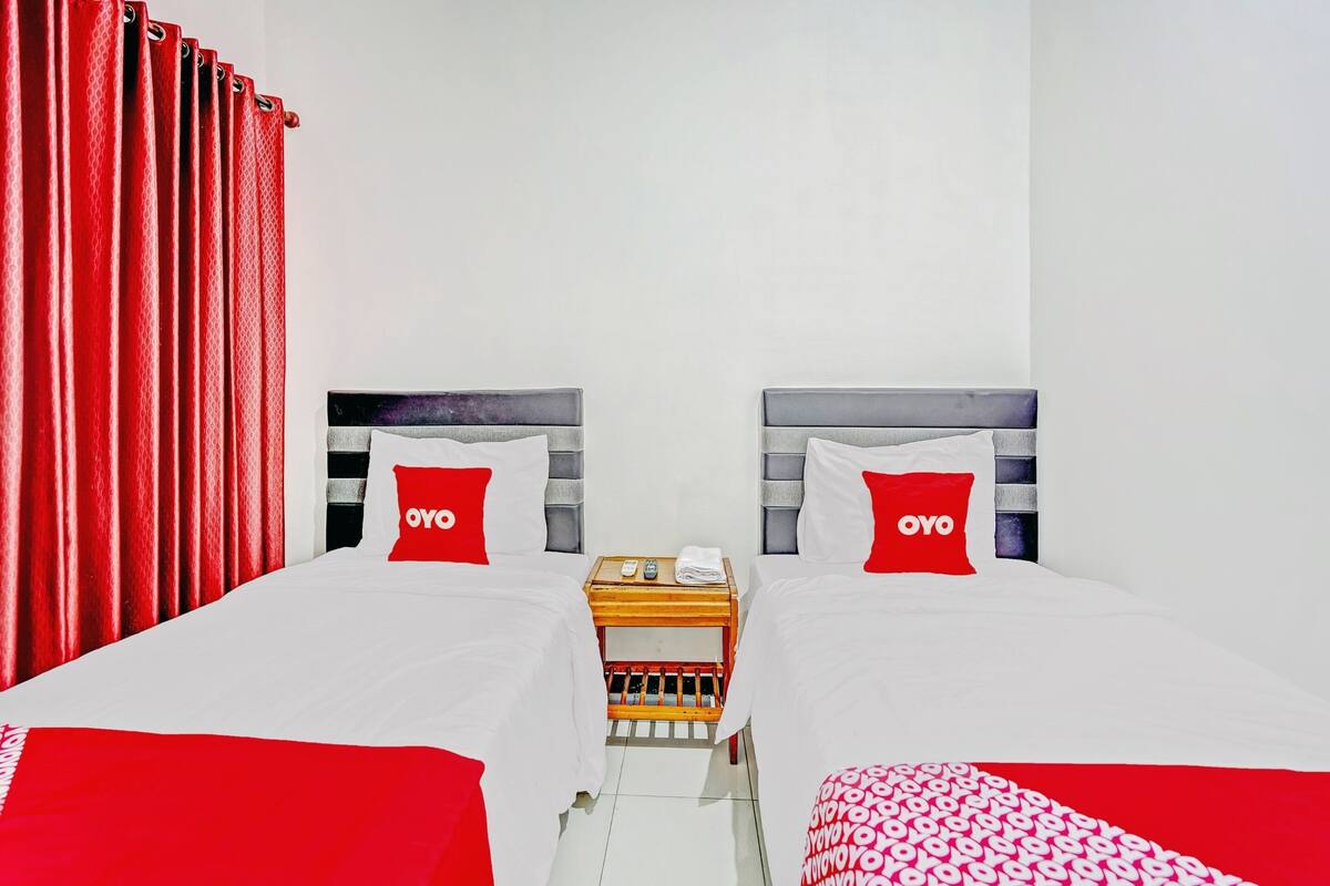 A hotel room with two beds, each with a red pillow labeled "OYO," a wooden nightstand with a telephone and a folded towel, and a red curtain on the left.