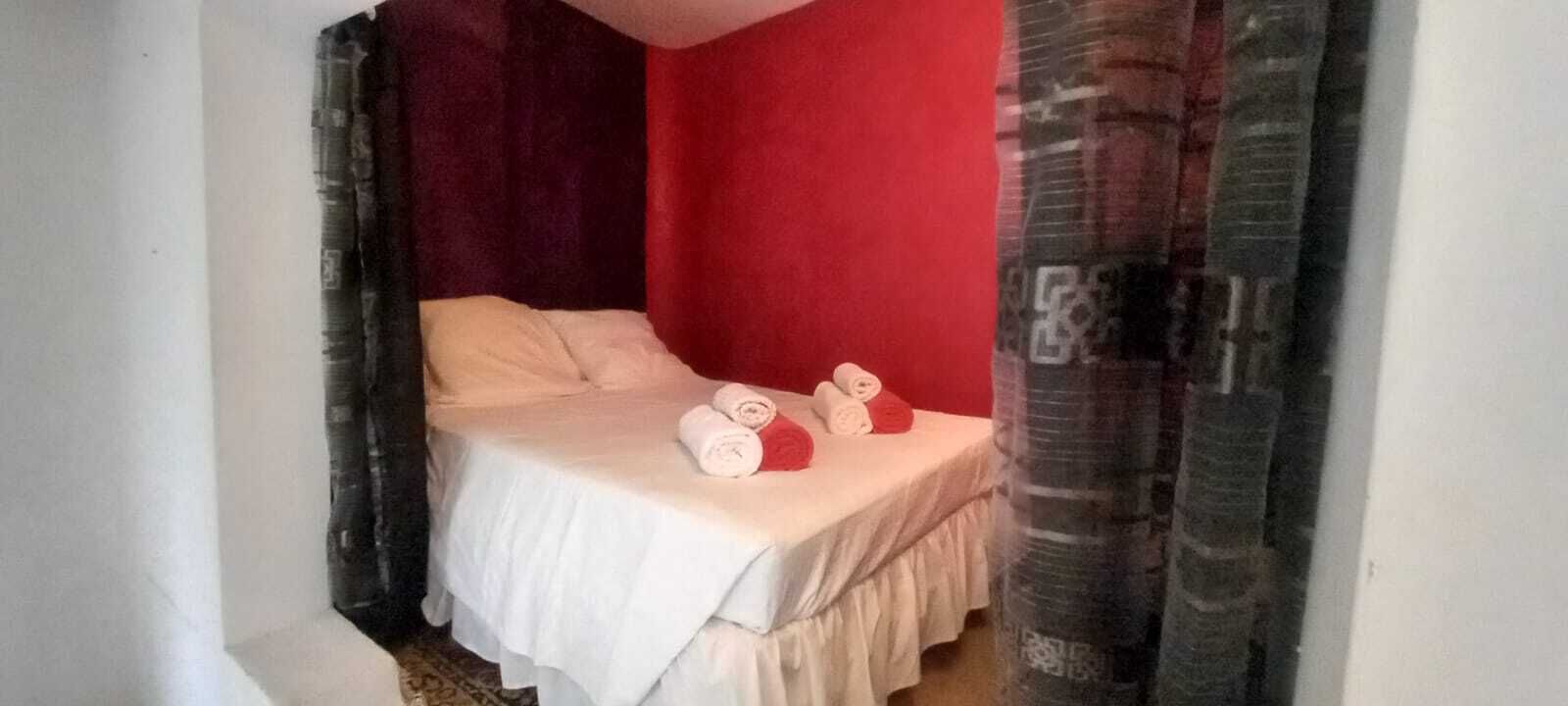 A single bed with a white bedspread, two rolled towels, and a red wall.