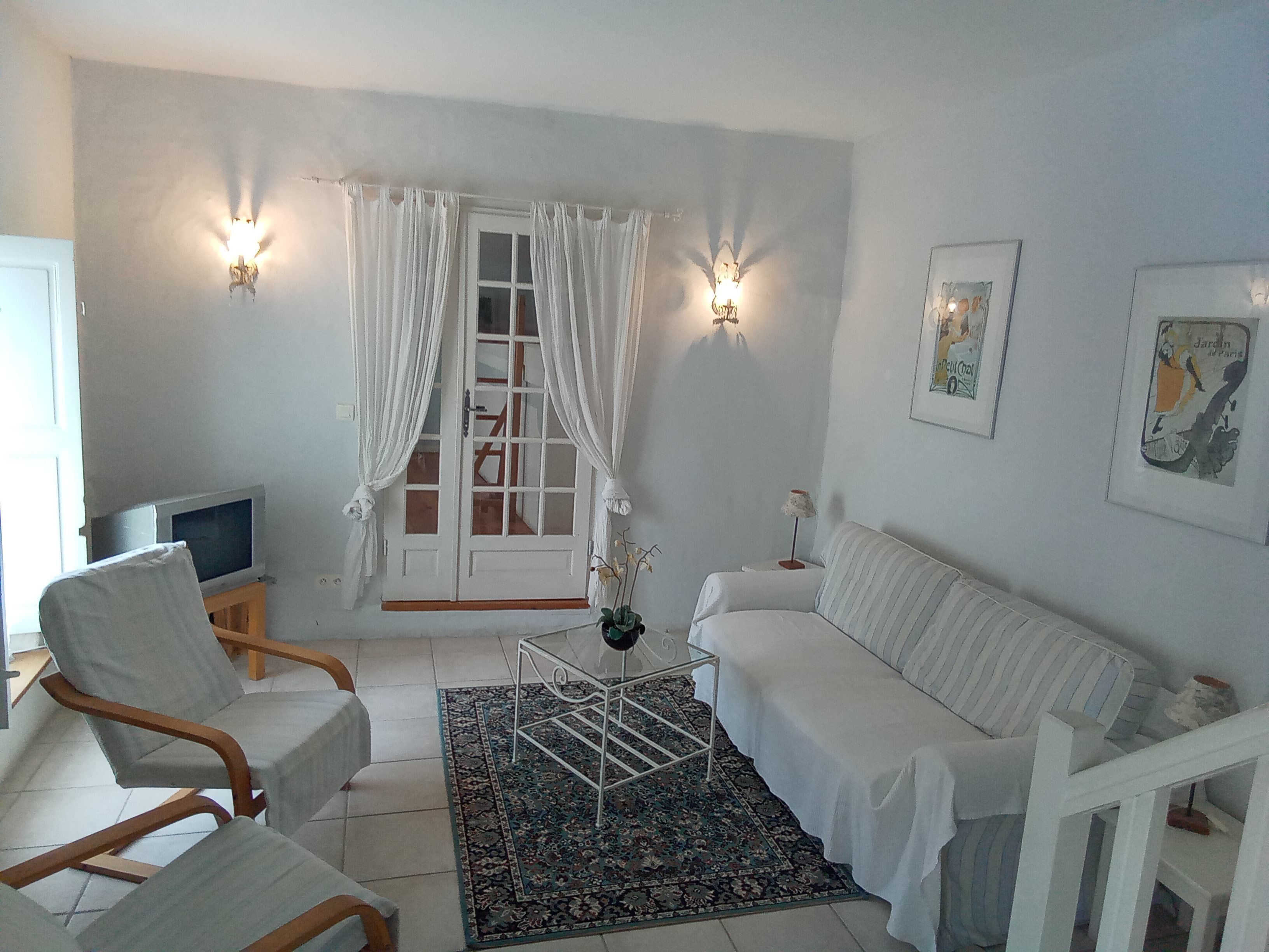 Una sala de estar con un sofá blanco, dos sillones, una mesa de centro, un televisor y un ventanal con cortinas.
